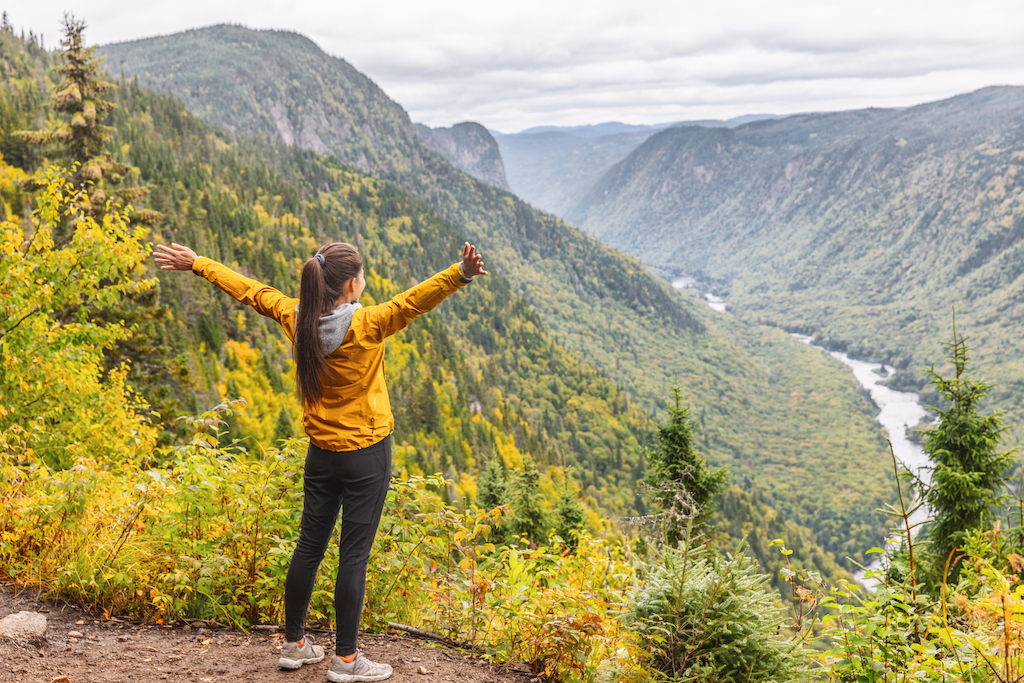 Happy woman hiking up mountain enjoying nature. Landscape with river view from top of trail hike. Girl with open arms outstretched in joy, enjoying travel fall in Jacques Cartier, Quebec, Canada.