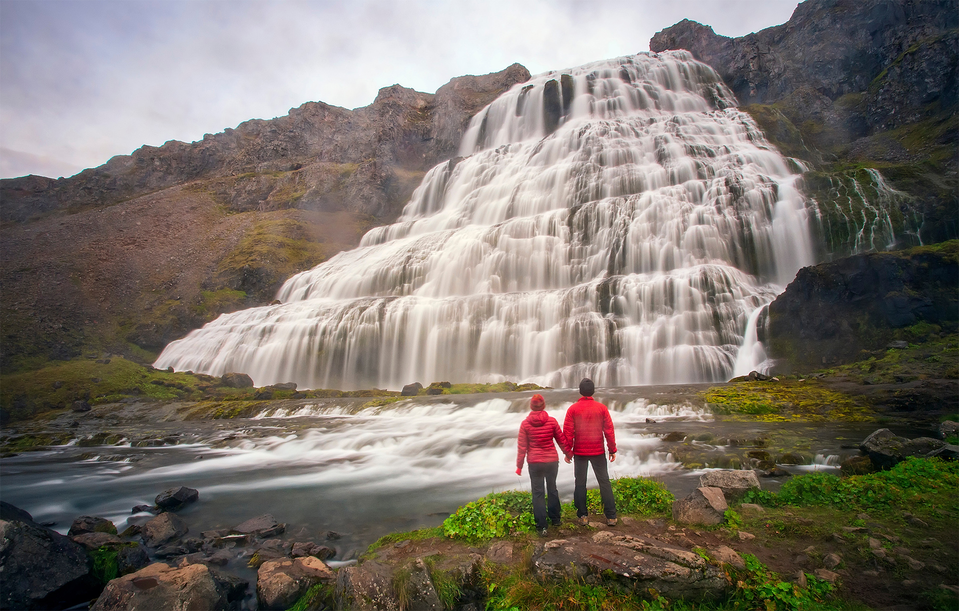 A couple holding hands at Dynjandi waterfall in the Westfjords of Iceland on a rainy day.