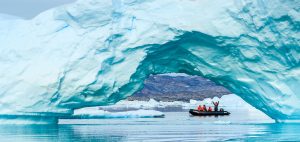 Travelers ride zodiac boat in arctic waters of Greenland underneath a giant iceberg arc