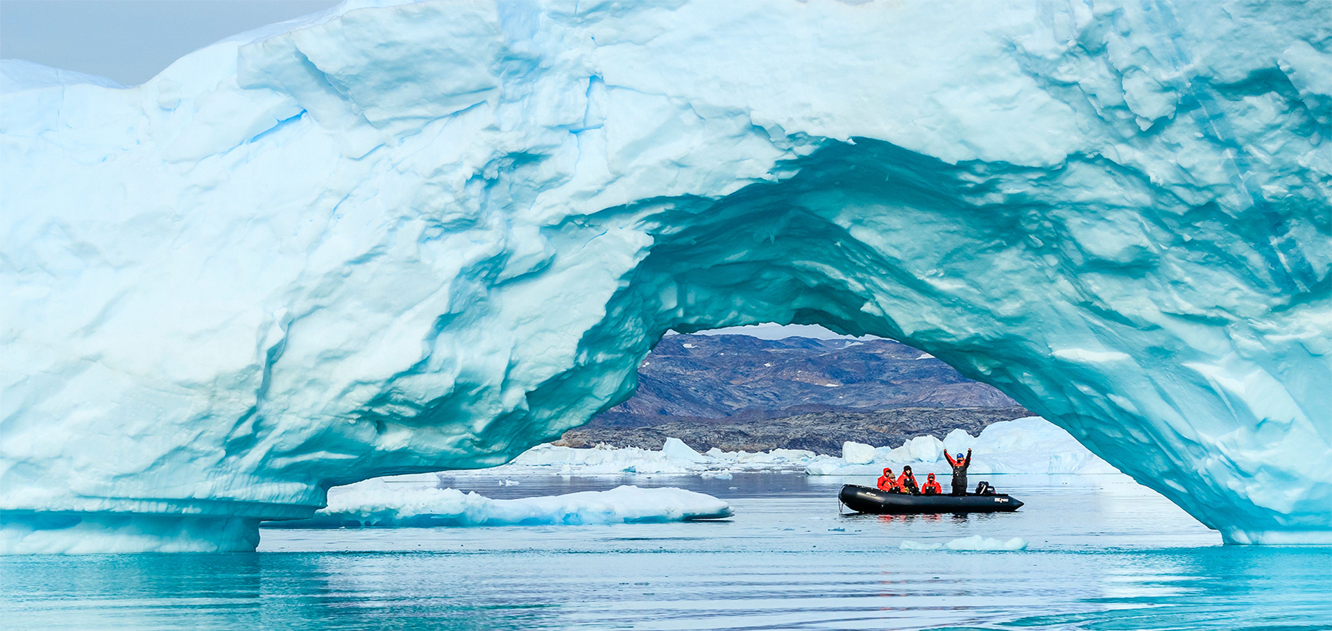 Travelers ride zodiac boat in arctic waters of Greenland underneath a giant iceberg arc