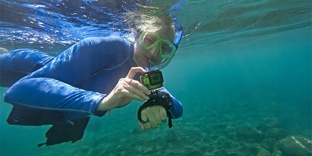 Judy's niece, Mary snorkeling underwater with a GoPro camera, wearing a blue rash guard and green snorkeling mask in clear, shallow water over a rocky seabed.